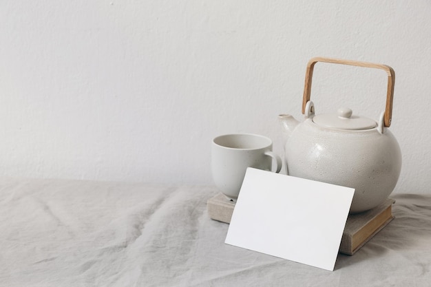Сцена завтрака натюрморт чашка кофе керамический чайник на старой книге пустая поздравительная открытка макет приглашения бежевая льняная скатерть белый фон стенынейтральная композиция утреннего образа жизни