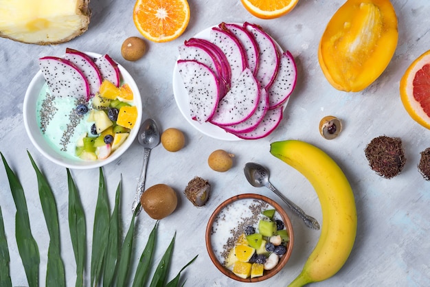 Photo breakfast table with yogurt acai bowls and fresh tropic fruits on a gray stone background with palm leaves, flat lay