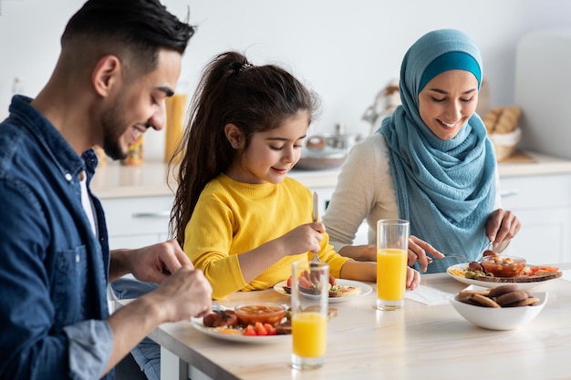 Photo breakfast time. middle eastern family of three enjoying tasty food in kitchen, happy arabic parents and their little daughter sitting at table, having delicious meal in the morning, closeup