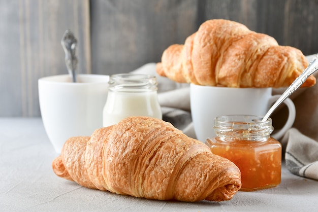 Breakfast two white cups of coffee with milk, two croissants and apple jam in a glass jar 