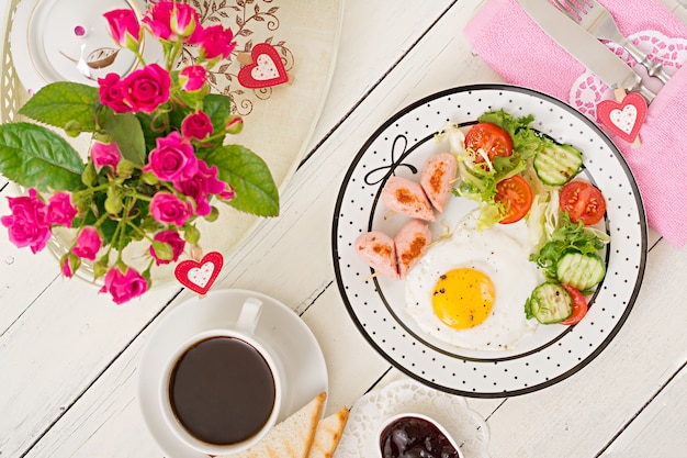 Breakfast on Valentine's Day - fried egg in the shape of a heart, toasts, sausage and fresh vegetables. Cup of coffee. English breakfast. Top view