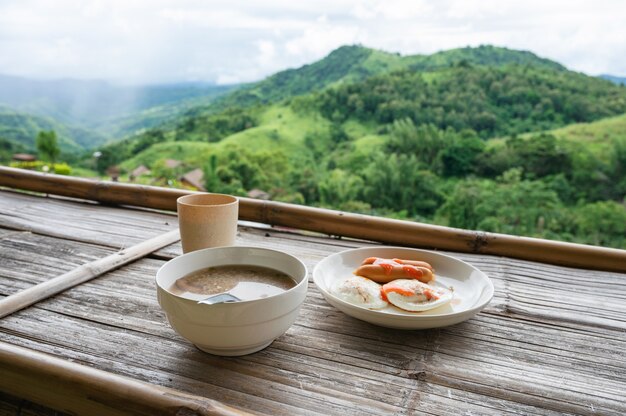 아침에 산의 전망을 감상할 수 있는 나무 카운터에 삶은 쌀, 소시지, 계란 프라이, 물이 포함된 아침 식사