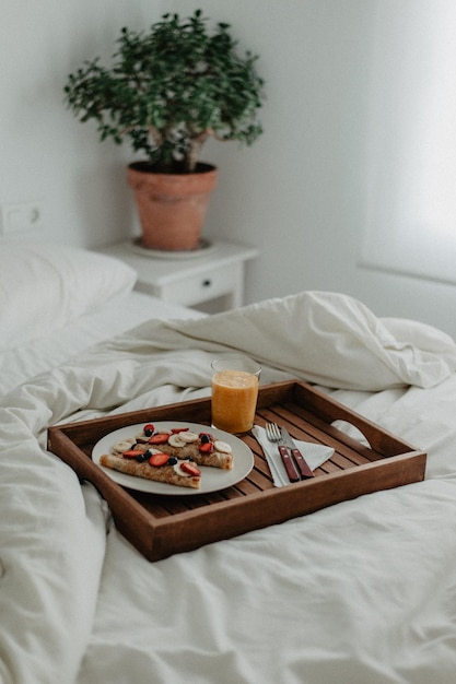 Breakfast with crepes, fruit and juice on wooden tray on white bed in a room.