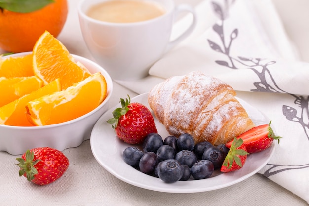 Breakfast with a cup of coffee, fresh croissant, strawberries and blueberries and juicy oranges