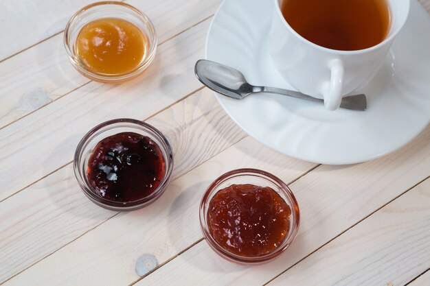 Colazione con una tazza di tè e gelatina di fragole, ribes e albicocche o marmellata su fondo bianco in legno rustico, primo piano