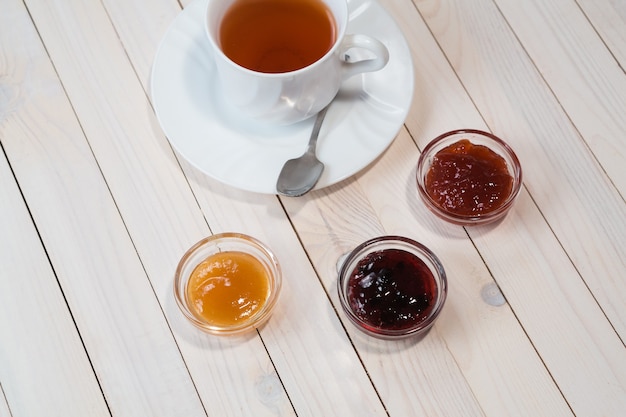 Breakfast with cup of tea and strawberry, currant and apricot jelly or jam on rustic wooden white background, close up