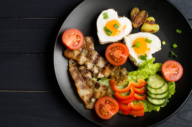 Breakfast with fried eggs, bacon and fresh vegetables. In a black plate, on a wooden background.