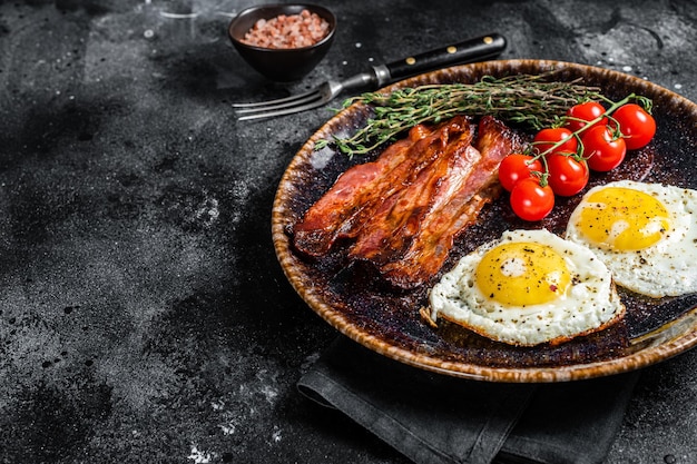 Breakfast with fried eggs and bacon with herbs in plate. Black background. Top view. Copy space.