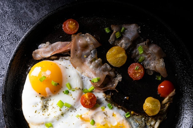 Breakfast with fried eggs in a pan with bacon and vegetables on dark background.