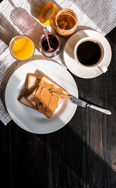 Завтрак с арахисовым маслом и вареньем и чашкой кофе на деревянном столе в лучах утреннего солнца