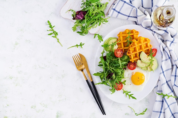 Breakfast with pumpkin waffles, fried egg, tomato, avocado and arugula on white surface