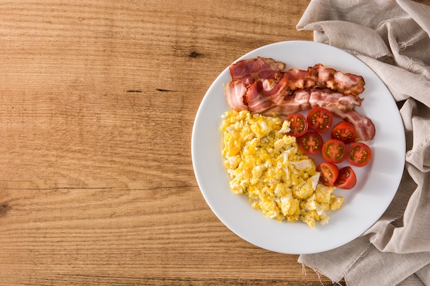 Breakfast with scrambled eggs, bacon and tomatoes on wooden table top view copy space