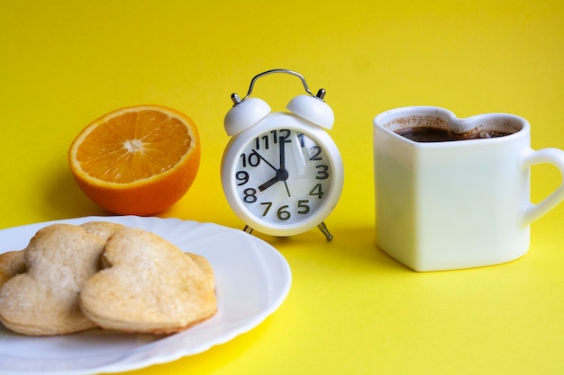 Завтрак на желтом столе, половина апельсина, кофе, печенье на белой тарелке и будильник