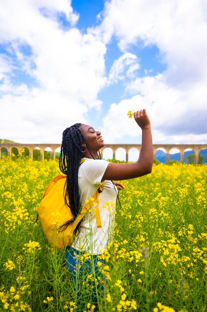 新鮮な空気と花の香りを呼吸する黄色い花のフィールドで旅行者を編んだ黒い民族の女の子