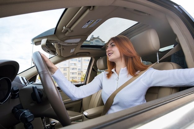 Brede hoekmening van jonge redhead vrouwenbestuurder die door veiligheidsgordel wordt vastgemaakt die een auto drijft die gelukkig glimlacht.