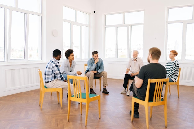 Brede opname van verschillende mensen van verschillende leeftijden die in stoelen zitten tijdens een ontmoeting met een psycholoog in het kantoor en problemen bespreken