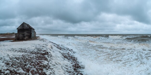 Breed panoramisch uitzicht op een dramatisch zeegezicht met een woeste witte zee en een vissershut aan de kust