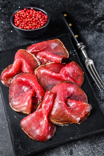 Bresaola cured meat beef cut pieces, Italian Antipasti. Black background. Top view.