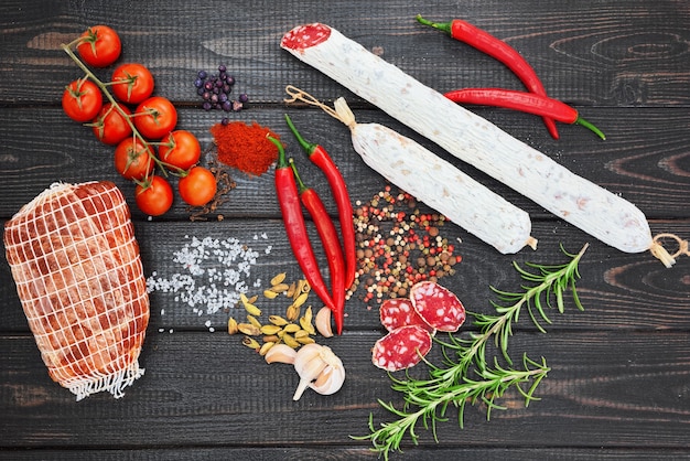 Bresaola and sliced cured sausage with spices, chili pepper, garlic and tomatoes on dark wooden rustic background.