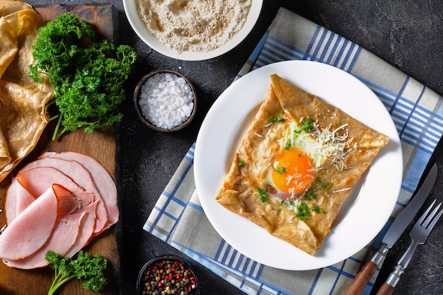 Breton crepe, Savory Buckwheat Galettes Bretonnes with fried egg, cheese, ham on a white plate on a kitchen table with ingredients on a cutting board, view from above, flatlay