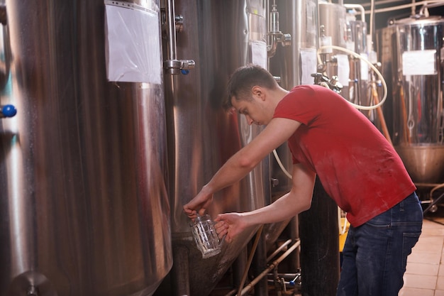 Photo brewery worker pouring fresh beer from metal tank into a glass