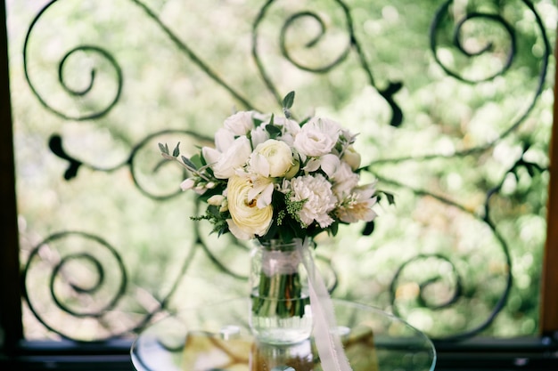 Photo bridal bouquet stands in a glass jar near the forged fence on the balcony