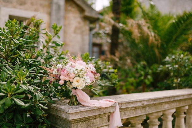 Bridal bouquet of white and pink roses peonies delphinium astilbe and white ribbons on the railing