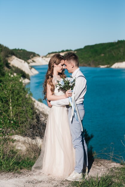 The bride in a beautiful dress hugging the groom in a light suit near the lake. Wedding couple standing on a sandy hill in the open air. A romantic love story. Azure-blue water on the horizon.