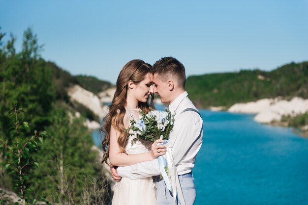 The bride in a beautiful dress hugging the groom in a light suit near the lake. Wedding couple standing on a sandy hill in the open air. A romantic love story. Azure-blue water on the horizon.