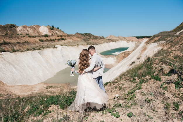 Foto la sposa in un bellissimo vestito che abbraccia lo sposo in una tuta leggera vicino al lago. coppie di cerimonia nuziale che si levano in piedi su una collina sabbiosa all'aperto. una romantica storia d'amore. acqua azzurro-azzurro all'orizzonte.