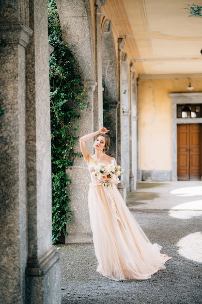 Bride in a beautiful dress with a bouquet of pink flowers stands in the vaulted hall with her hand