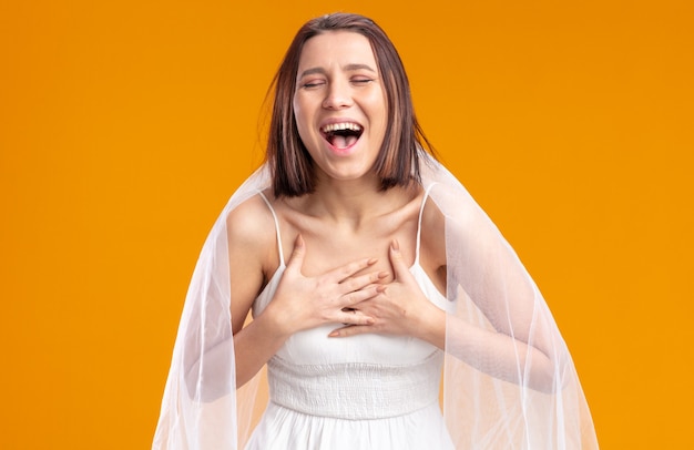 Sposa in un bellissimo abito da sposa felice ed emozionata che ride con le mani sul petto in piedi sul muro arancione