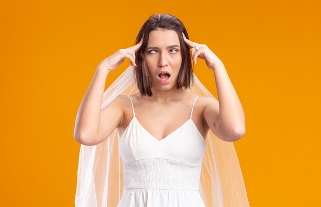 Bride in beautiful wedding dress looking annoyed rolling eyes up touching her temples with index fingers standing over orange wall