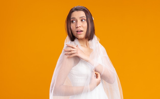 Bride in beautiful wedding dress with veil looking aside happy and surprised standing on orange