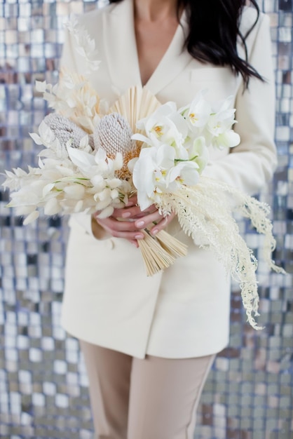 Bride in beige suit holds wedding boho bouquet with orchids banksia and dry flowers on a silver sequin texture background