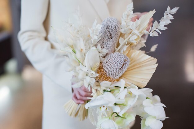 Невеста в бежевом костюме держит свадебный букет в стиле бохо с орхидеями банксия и сухими цветами