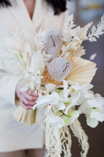 Bride in beige suit holds wedding boho bouquet with orchids banksia and dry flowers