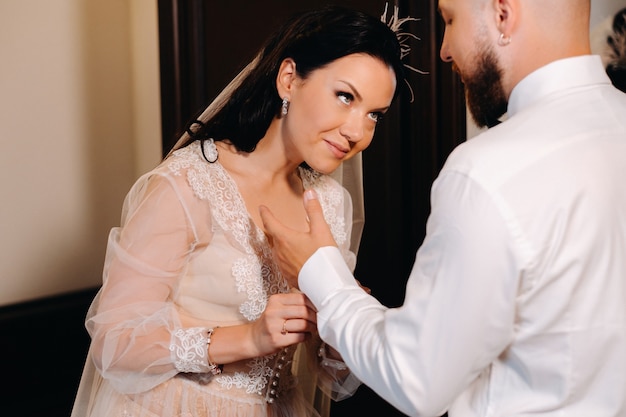 Невеста в будуарном нижнем белье наряжает жениха в интерьер гостиницы.