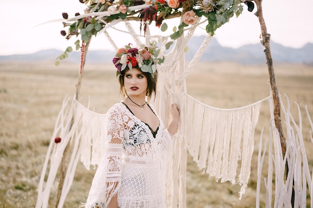 Bride in a braided dress and a wreath stands near the wedding arch in the field