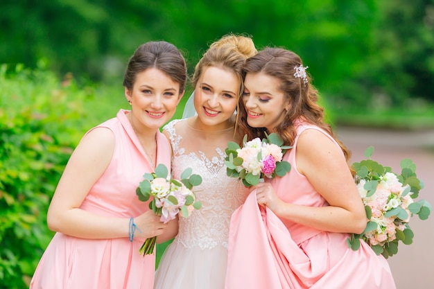 La sposa e le damigelle in abiti rosa si divertono e posano nel parco. ragazze che tengono i mazzi di nozze.