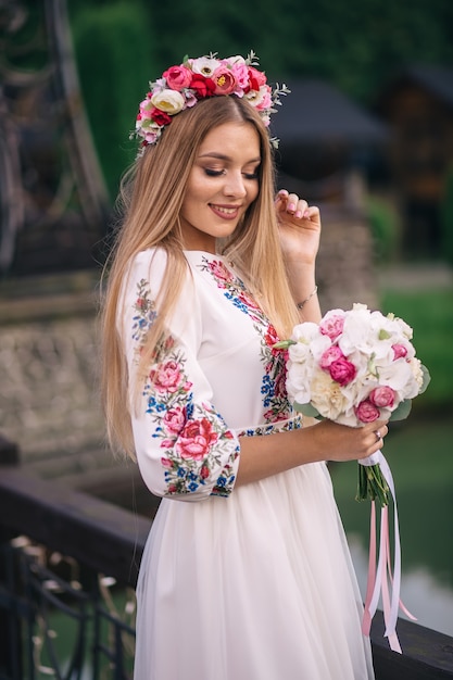 Bride in chic embroidered dress with wreath on her head looks at wedding bouquet of roses.