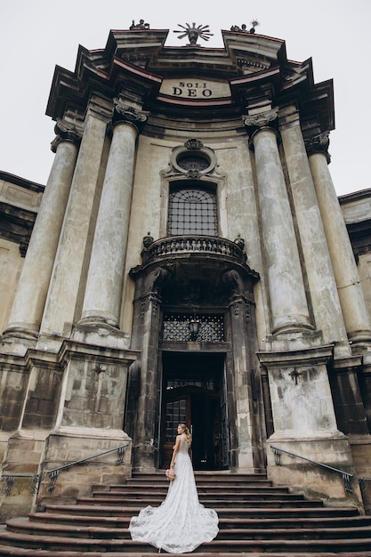 Невеста в платье с длинным шлейфом стоит перед старой готической церковью