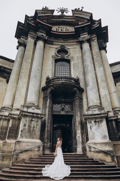 Bride in dress with long train stands before an old Gothic church