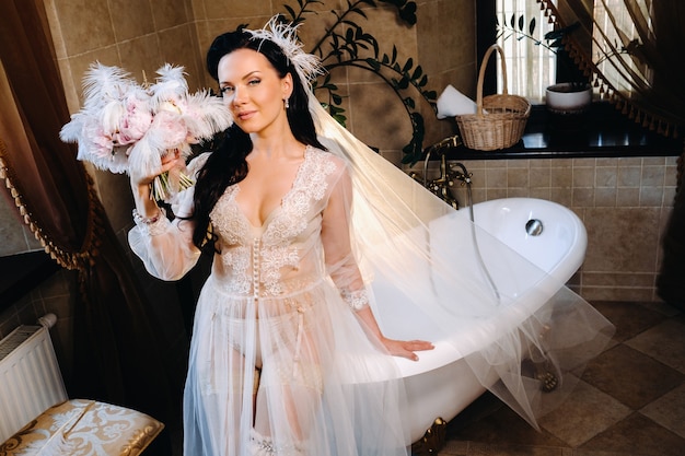 The bride, dressed in a boudoir transparent dress and underwear, sits near a vintage bath with a feather in her hands, The morning of the bride.