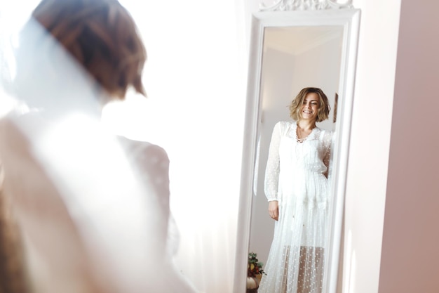 Photo bride in elegant white dress looking in mirror the bride smiles and poses in front of the mirror