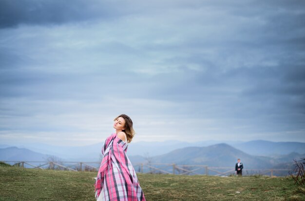 Bride envlopes herself in pink plaid posing on green hill in coudy day