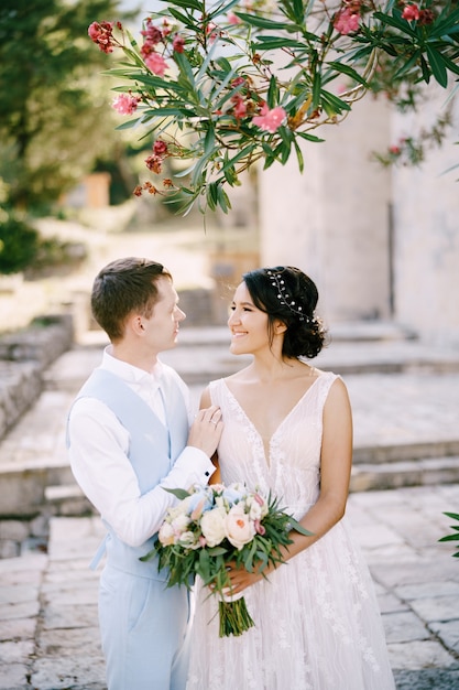 The bride and groom are embracing under a blooming oleander the bride is holding a bouquet in her