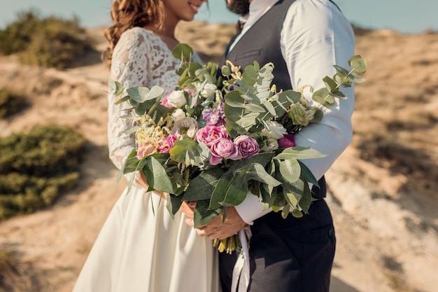 The bride and groom are holding a bouquet of pink flowers and greenery in the background mountains