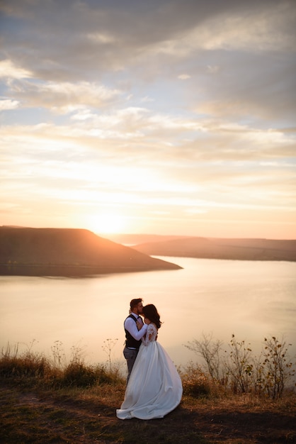 La sposa e lo sposo si abbracciano sullo sfondo del lago durante il tramonto