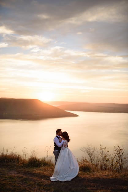 The bride and groom are hugging on the background of the lake during sunset